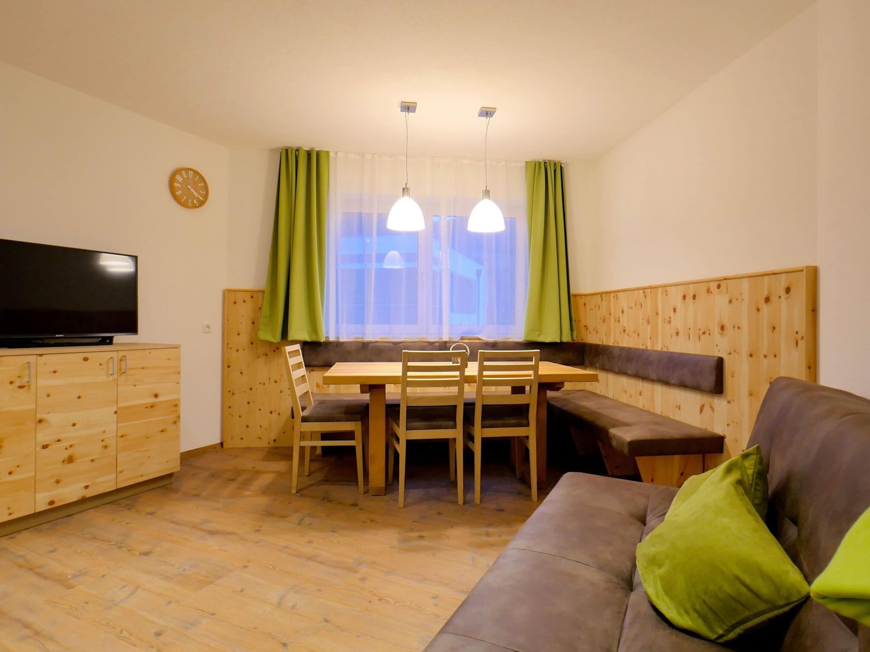 A cozy room with a wooden dining table, chairs, a sofa, a television, and a window with curtains.