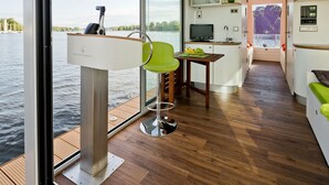 Un interior moderno con una terraza de madera, una silla verde, una mesita con frutas, un televisor y un lavabo.