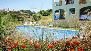 Une piscine entourée de fleurs rouges et de verdure, avec un bâtiment doté de balcons et d’un espace aménagé au bord de la piscine.