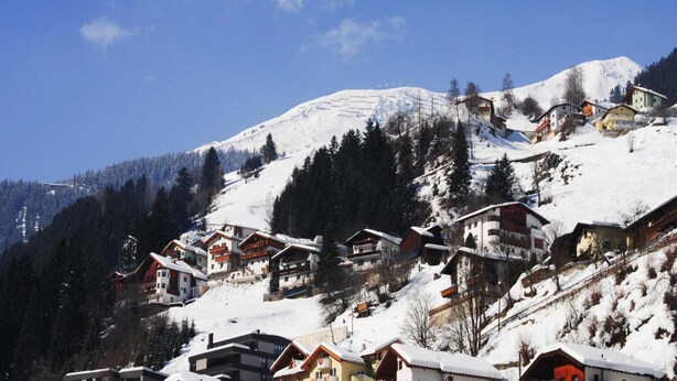 Un village de montagne enneigé, avec ses maisons traditionnelles en bois, un ciel bleu azur et un sommet enneigé imposant qui se détache à l’arrière-plan.