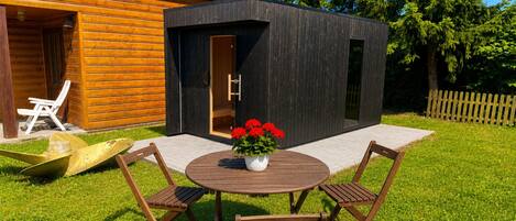 A wooden cabin with a modern extension, a round wooden table with chairs, and a potted plant with red flowers.