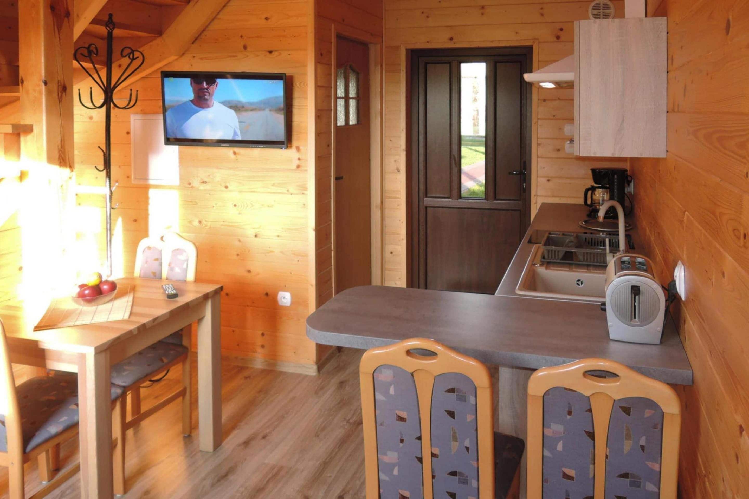 A wooden cabin interior with a dining table, chairs, a television mounted on the wall, a kitchen area with a sink and a coffee maker, and a door leading outside.