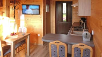 A wooden cabin interior with a dining table, chairs, a television mounted on the wall, a kitchen area with a sink and a coffee maker, and a door leading outside.