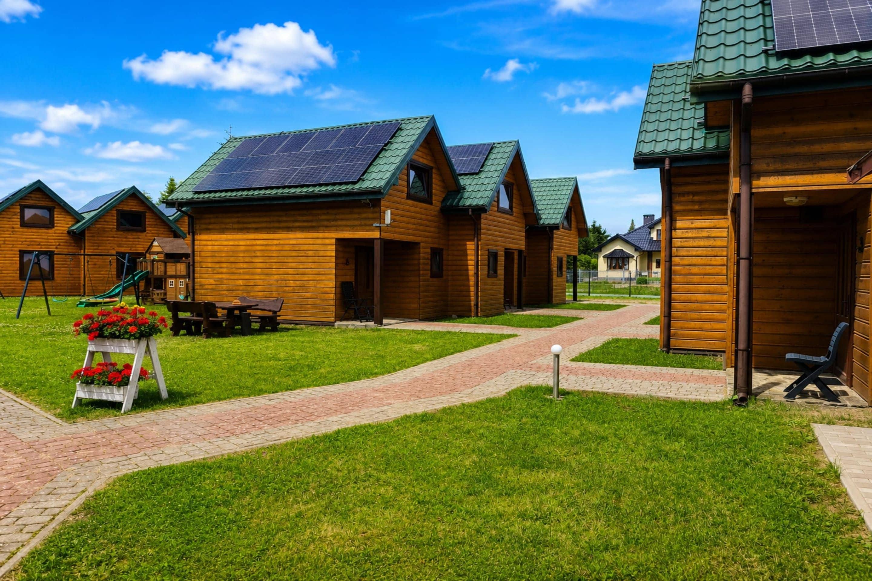 A row of wooden cabins with green roofs and solar panels, a paved walkway, and a small garden area with flowers.