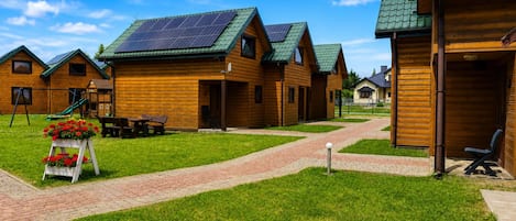 A row of wooden cabins with green roofs and solar panels, a paved walkway, and a small garden area with flowers.