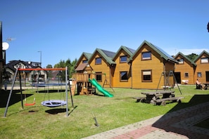 A row of wooden cabins with pitched roofs, a playground with a slide, and picnic tables. - Terraced House in Rewal Near Sandy Beach (Rewal)