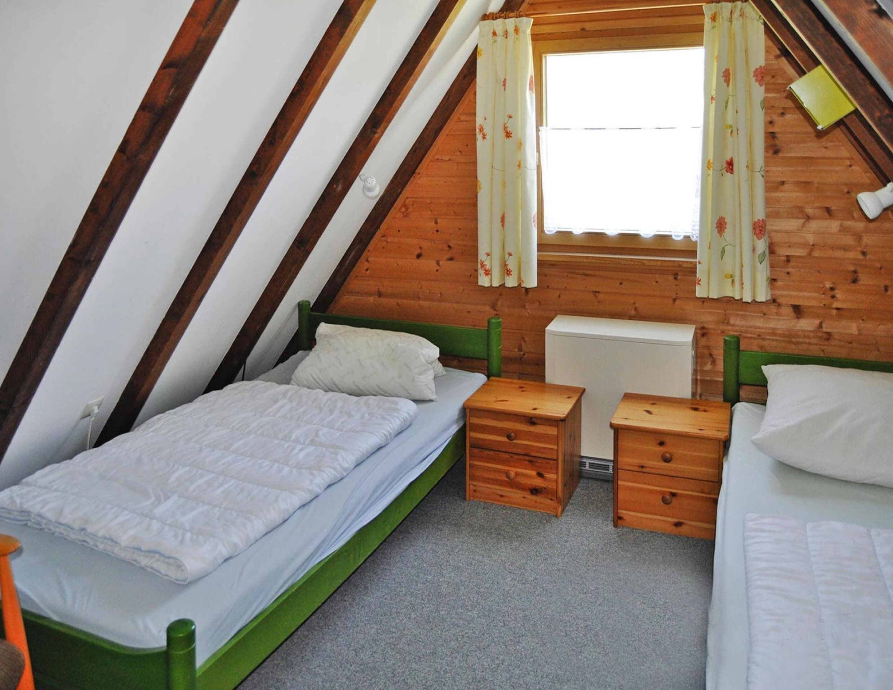 A small, cozy attic bedroom with two single beds, wooden nightstands, and a window with floral curtains.
