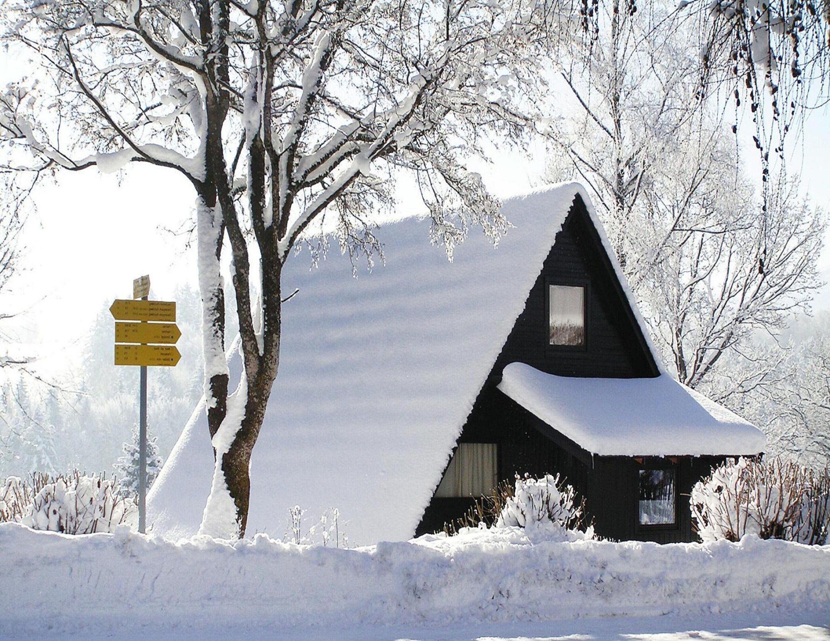 A snow-covered cabin with a triangular roof, surrounded by snow-laden trees.