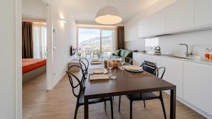 A modern kitchen with white cabinets, a dining area with a table set for two, and a view of a mountain landscape through large windows. - Residence Olivo, Garda (Garda)