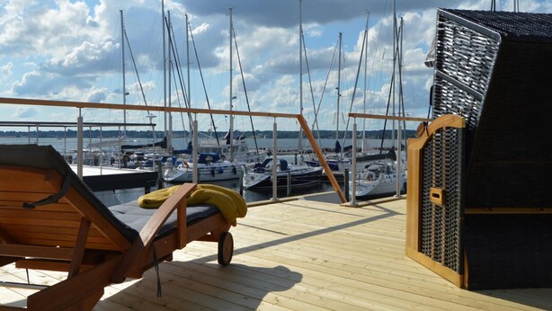 Een houten terras met een ligstoel en een klein houten gebouwtje, met uitzicht op een jachthaven vol boten.