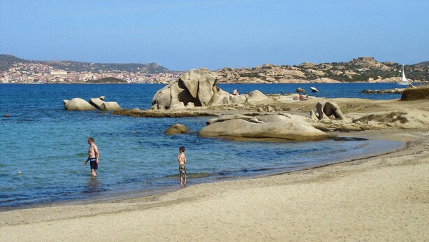 Una playa con rocas grandes en el agua, una orilla arenosa y un paisaje urbano distante.