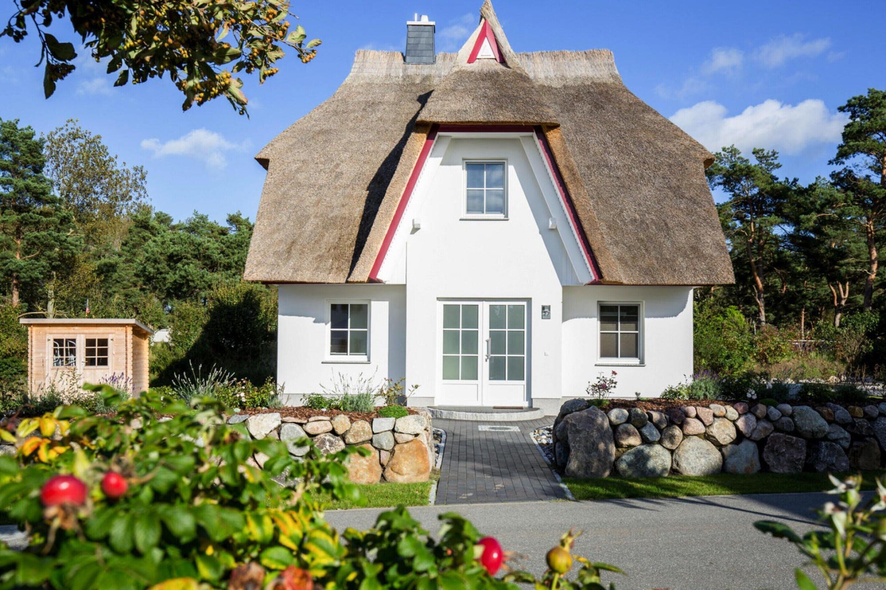 A white house with a thatched roof, a stone wall, and a small wooden structure in the background.