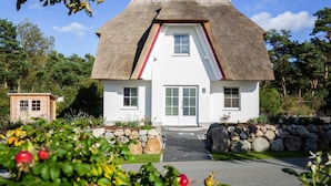 A white house with a thatched roof, a stone wall, and a small wooden structure in the background. - Ferienhaus, Fuhlendorf (Fuhlendorf)
