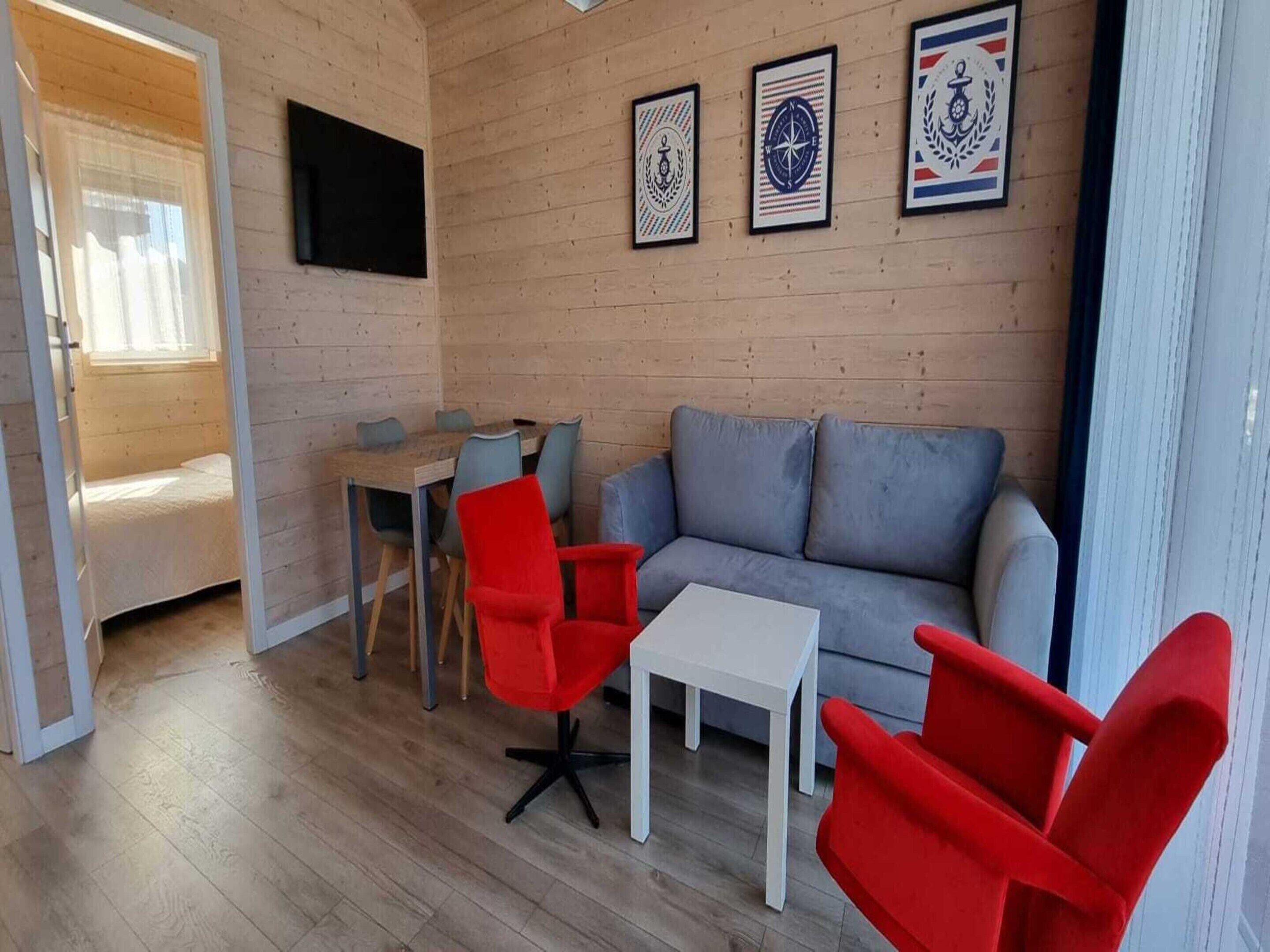 A cozy living room with a grey sofa, red chairs, a white coffee table, and a wooden dining table. There are framed pictures on the wall and a TV mounted above the sofa.
