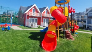 A playground with a slide and climbing structures, surrounded by a fenced area and residential buildings. - Single-story Holiday Homes, Pool, Playground (Mielno)