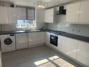 A modern kitchen with white cabinetry, a built-in washer and dishwasher, a gas stove, and a window with blinds. - 10min to Oxford - 3 Bedrooms - Sleeps 7 - Pets Ok (Kidlington)