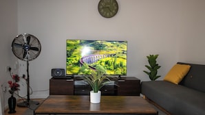 Ein Wohnzimmer mit Flachbildfernseher, Standventilator, Topfpflanze, Sofa und einer Wanduhr.