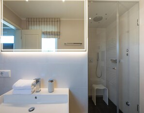 A modern bathroom with a white sink, a mirror, a shower, and a small stool. - Appartement in Fantastischer Lage auf Nordstrand (Nordstrand)