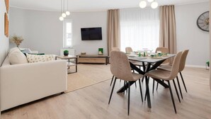 A modern living room with a sofa, dining table, and chairs. There is a flat-screen TV mounted on the wall and a window with curtains. - Spacious Apartment,summer Swimming Pool, Pobierowo (Rewal)