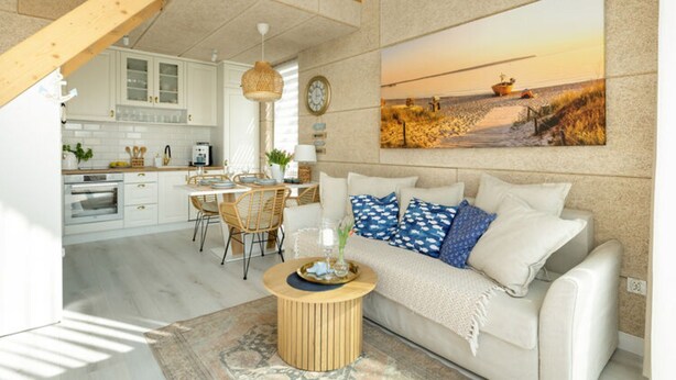 A modern kitchen with white cabinets, a dining area with a wooden table and chairs, and a living room with a beige sofa and a painting of a beach scene. - Comfortable House With, Whirlpool Bathtub Wrzosowo (Kamien Pomorski)