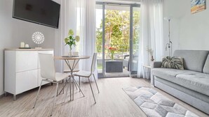 A modern living room with a grey sofa, a dining table with chairs, a TV mounted on the wall, and a balcony with a view of trees and a building. - Apartment in Pobierowo Near Baltic Beach (Rewal)