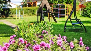 Un’area giochi con una casetta in legno e uno scivolo, circondata da erba verde e fiori viola.