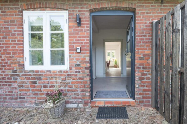 A brick building with a white-framed window, a wooden door, and a small basket with flowers. - Stylische Ferienwohnung in Landlicher Idylle (Wobbenbüll)