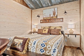 A wooden-themed bedroom with a bed, pillows, a nightstand, wall art, and a sign reading 'TEXAS RALEIGHS.' - Rockin' R - Shooting Star Cabin (Fredericksburg)