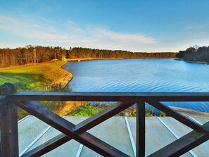 A view from a covered deck overlooking a calm lake with a forested shoreline. - Holiday Home, Private Garden and Lake Access (gmina Manowo)