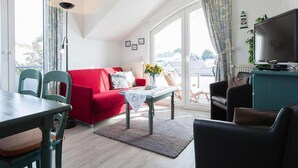 Un salon avec un canapé rouge, une table à manger avec des chaises, un téléviseur posé sur un meuble et une vue sur l’extérieur grâce à de grandes fenêtres.
