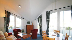 Un salon comprenant un canapé, une table à manger avec des chaises, un téléviseur posé sur un meuble et de grandes fenêtres avec des rideaux.