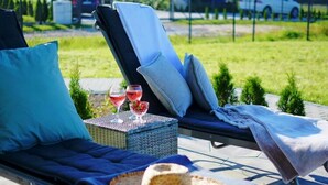Une chaise longue avec un panier tressé et un chapeau, une petite table avec des verres de vin rouge, et une pelouse verdoyante en arrière-plan.