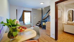 Un intérieur moderne avec un coin repas comprenant une table en bois, des chaises et un vase orné de fleurs et de fruits. On aperçoit un escalier au fond, menant à un niveau supérieur.
