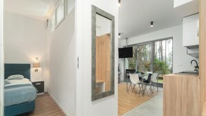 A modern hotel room with a bed, bedside table, lamp, and a large mirror. There is a dining area with a table, chairs, and a TV. The room has large windows and a view of trees. - Comfortable Apartment Close to a Sandy Beach (Rewal)