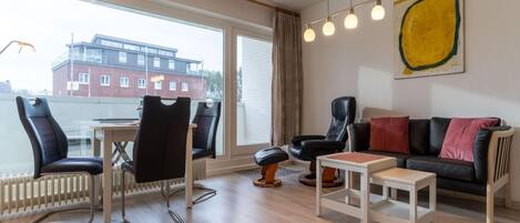 Un salon moderne avec vue sur un immeuble, grâce à de grandes fenêtres, comprenant un canapé en cuir noir, une table basse blanche et un fauteuil en cuir noir.