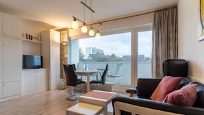 Ein modernes Wohnzimmer mit einer Couch, einem Esstisch und einem Fernseher. Ein Balkon bietet Blick auf Gebäude und Bäume.