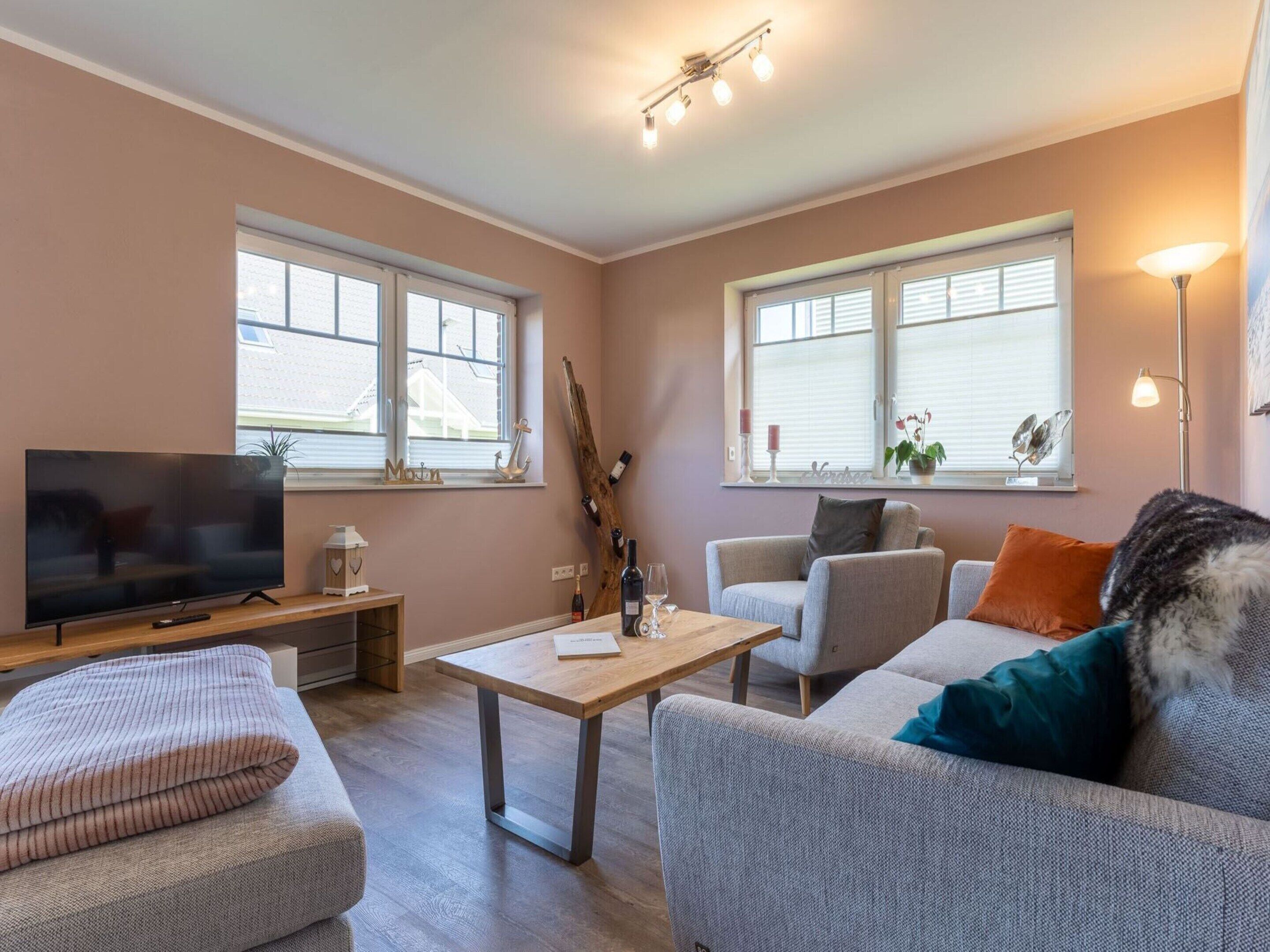 A modern living room with a flat-screen TV, a wooden coffee table, grey sofas, and a large window with blinds.