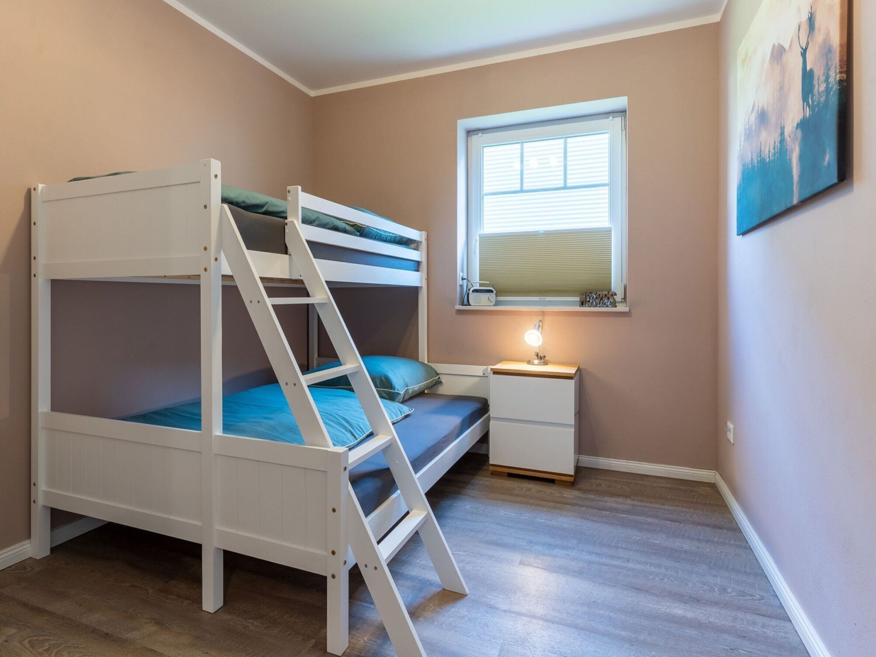 Uma cama beliche com escada, uma mesinha de cabeceira com luminária, uma janela com persianas e um quadro na parede.