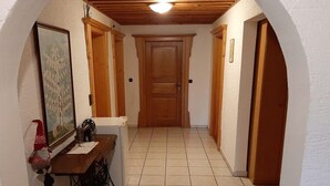 Un couloir se termine par une porte en bois, une machine à coudre est posée sur une table, et un tableau encadré orne le mur.