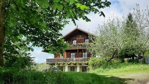 Une maison en bois au style rustique, dotée d’un balcon, entourée d’une végétation luxuriante et surplombant un ciel dégagé.