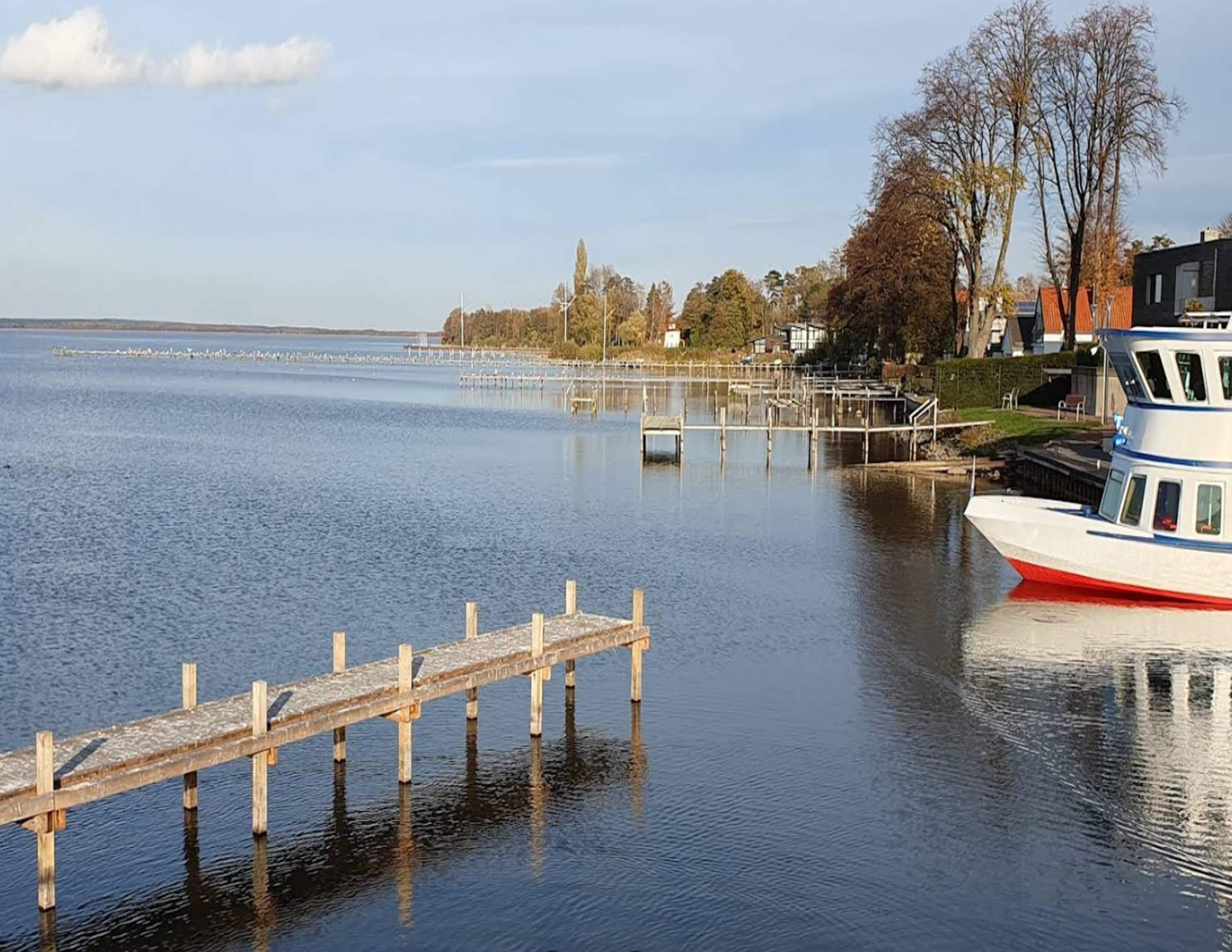 Un lago tranquilo con un muelle de madera que se adentra en el agua, una embarcación blanca con detalles rojos amarrada al muelle, y una hilera de casas al fondo.