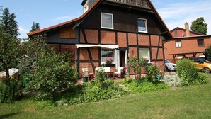 Una casa a due piani con tetto ripido, un balcone e un piccolo giardino con piante in vaso e una panchina.