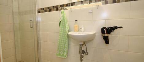 A bathroom with a glass shower enclosure, a sink with a mirror above it, and a hairdryer mounted on the wall.