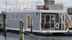 Una casa flotante anclada en un muelle, con terraza techada, ventanas y un flotador salvavidas.