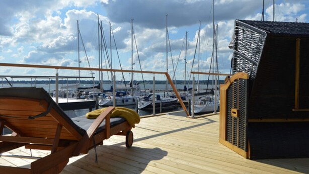 Una terraza de madera con una silla de descanso y un edificio pequeño, con vista a un puerto deportivo con varias embarcaciones.