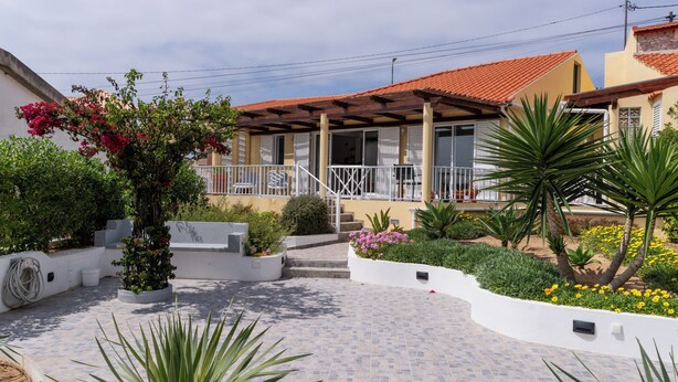 Um edifício residencial com varanda coberta, um jardim com diversas plantas e flores, e um espaço de pátio com piso de azulejos.