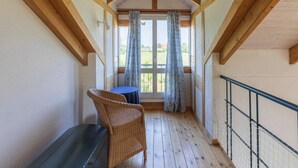 A covered balcony with wooden beams, a wicker chair, a table, and a view of a green field through large windows with patterned curtains. - Liebevoll Eingerichtetes Ferienhaus in Langenhorn (Langenhorn)