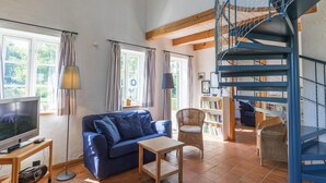 Un salon avec un canapé bleu, des fauteuils en osier, une petite table, un téléviseur et une bibliothèque.