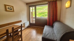 A room with a wooden table and chairs, a grey sofa, a window with red curtains, and a wall-mounted light fixture.
