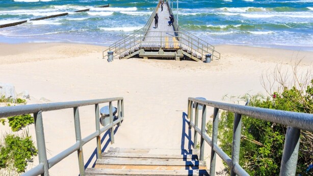 Una pasarela de madera que conduce a una torre de salvavidas en una playa, con olas al fondo.